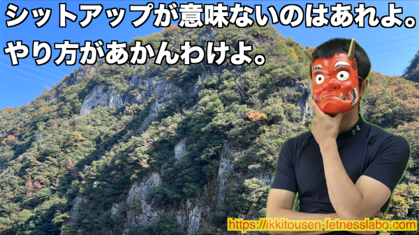 　赤鬼のお面をつけた男性が「シットアップが意味ないのはやり方が悪いから」と背景の山を背に語っている様子